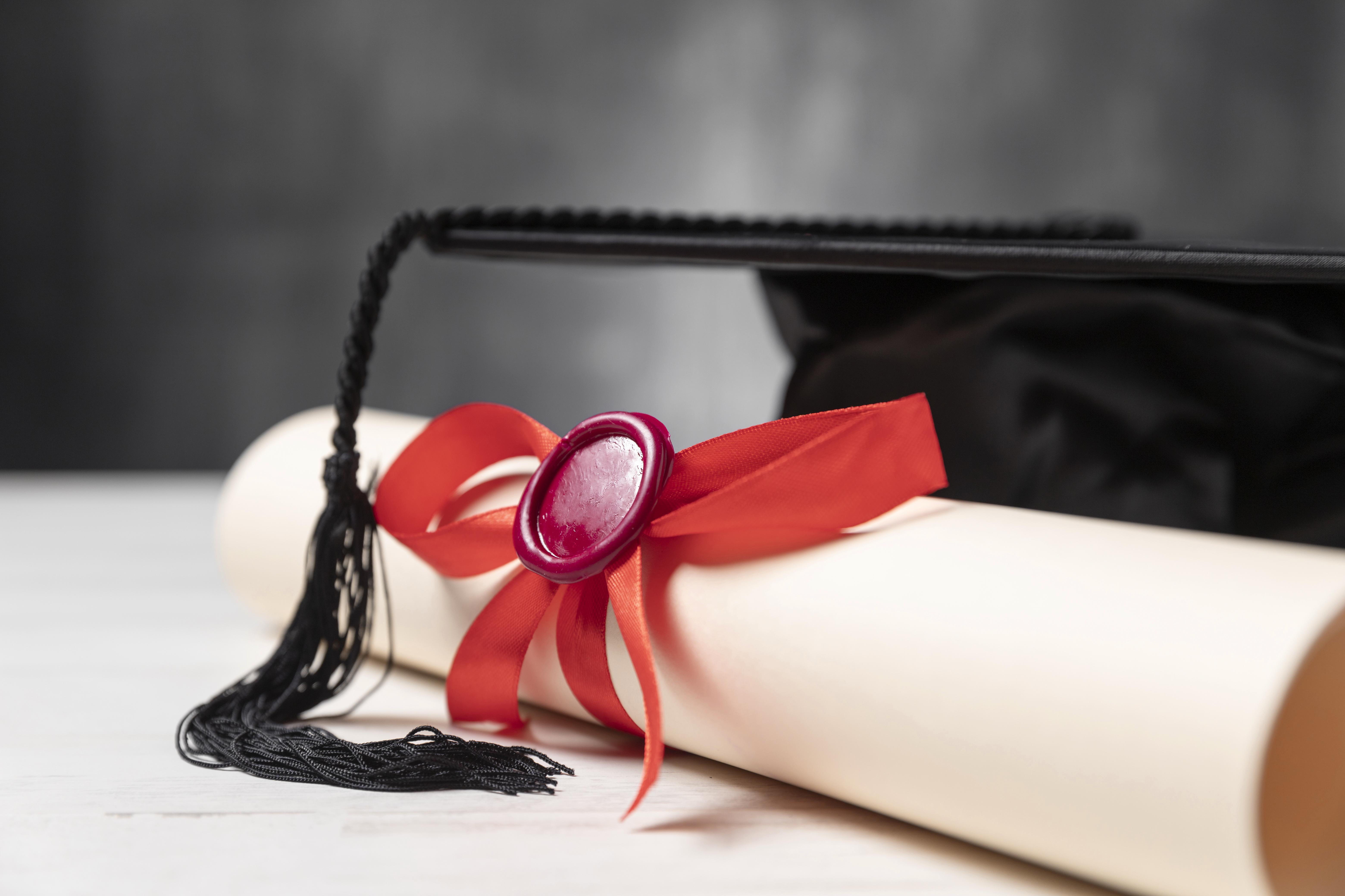 graduation diploma shown sealed with a red seal and bow, a graduation cap sits beside the scroll with the tassel on the scroll